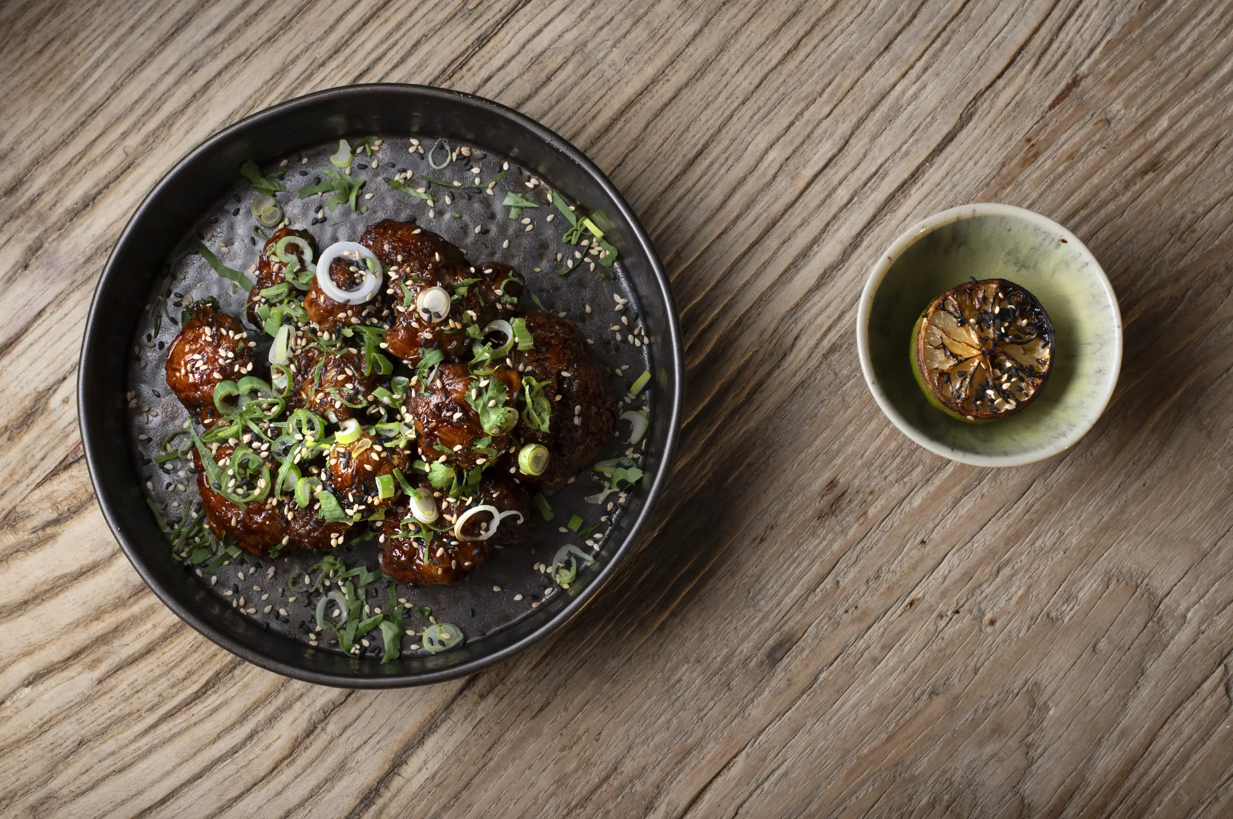 A glazed dish with spring onion and sesame beside a charred lime, on the oak table at Saskia's